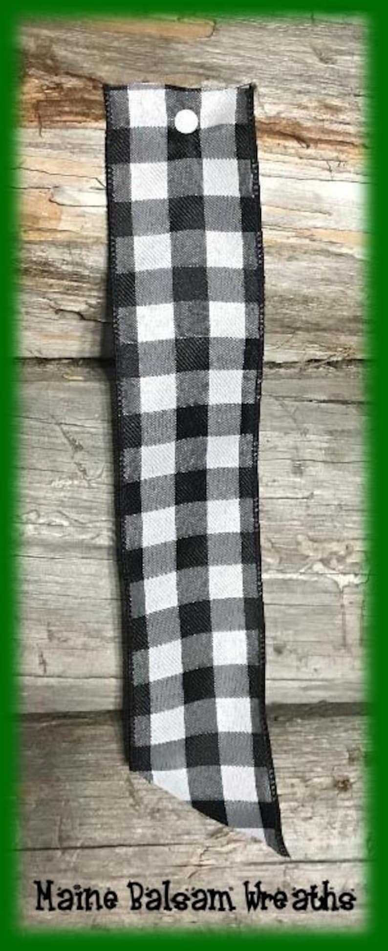 May include: A black and white checkered ribbon with a white button at the top. The ribbon is displayed vertically against a wooden background. The text "Maine Balsam Wreaths" is at the bottom.