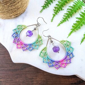 May include: A pair of handmade macrame earrings with a rainbow gradient design. The earrings feature a half-circle shape with intricate knotting, transitioning from green to pink and blue. Each earring has a silver hook and a faceted purple gemstone pendant.