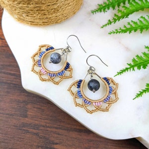 Op de afbeelding: Een paar handgemaakte oorbellen met een delicaat mandala-ontwerp in een kleurenschema van blauw, roze en goud. Elke oorbel heeft een donkergrijze stenen kraal die in het midden hangt.