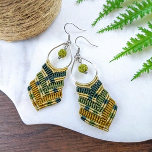 May include: A pair of green, gold, and black macrame earrings with lava rock beads. The earrings are hanging from silver hooks.