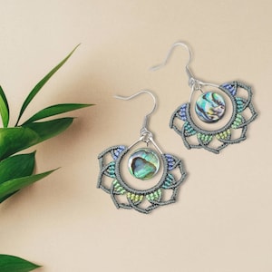 May include: A pair of handcrafted earrings featuring a fan-shaped design with a central abalone shell. The earrings have a silver-tone hook and are adorned with intricate macrame detailing in shades of gray, blue, and green.