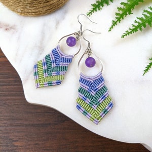 May include: A pair of handmade macrame earrings with purple gemstone beads. The earrings are made with green, blue, and purple cord and have a diamond shape.