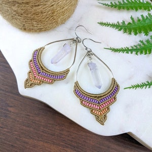 May include: A pair of dangle earrings with a teardrop-shaped design. The earrings are made with a macrame technique using brown, purple, and orange thread. Each earring features a clear quartz crystal pendant.