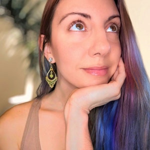 Puede incluir: Una mujer con el pelo largo y multicolor lleva un par de pendientes de oro con detalles verdes y negros. Los pendientes tienen forma de lágrima y presentan una pequeña piedra redonda verde.