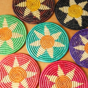 May include: Six round woven coasters with a starburst design in various colors, including green, blue, purple, red, and brown. The center of each coaster features a spiral design in orange.