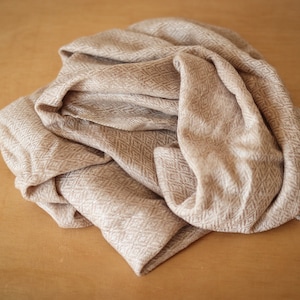 May include: A beige knitted scarf with a herringbone pattern. The scarf is folded and arranged on a wooden surface.