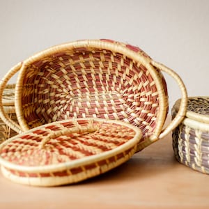 May include: Three woven baskets with lids. The largest basket is oval shaped and has a red and natural woven pattern. The two smaller baskets are round and have a natural and dark brown woven pattern.