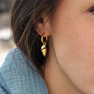 May include: A gold-colored hoop earring with a textured, leaf-shaped charm. The earring is worn on the ear of a person wearing a blue top. The earring is a simple, elegant piece of jewelry.