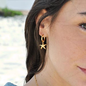 May include: Gold-colored hoop earring with a starfish charm. The earring is worn on the ear of a person with dark hair. A gold necklace is also visible. The background is blurred, with a hint of greenery and water.