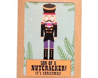 Puede incluir: Una tarjeta de Navidad con un soldado cascanueces de dibujos animados sobre un fondo azul claro con nieve cayendo y ramas de pino. La tarjeta dice "SON OF A NUTCRACKER! IT'S CHRISTMAS!" en un cuadro marrón, en un marco de tarjeta marrón.