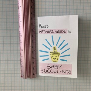 May include: A pink and white card with the title "Annie's Wayward Guide to Baby Succulents". The card features a cartoon illustration of a potted succulent with a smiling face.