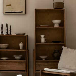May include: Two wooden bookshelves with open shelves and drawers, filled with ceramic bowls, vases, and other decorative items. A wicker basket sits on top of one shelf. A beige armchair with a cushion sits in front of the shelves.