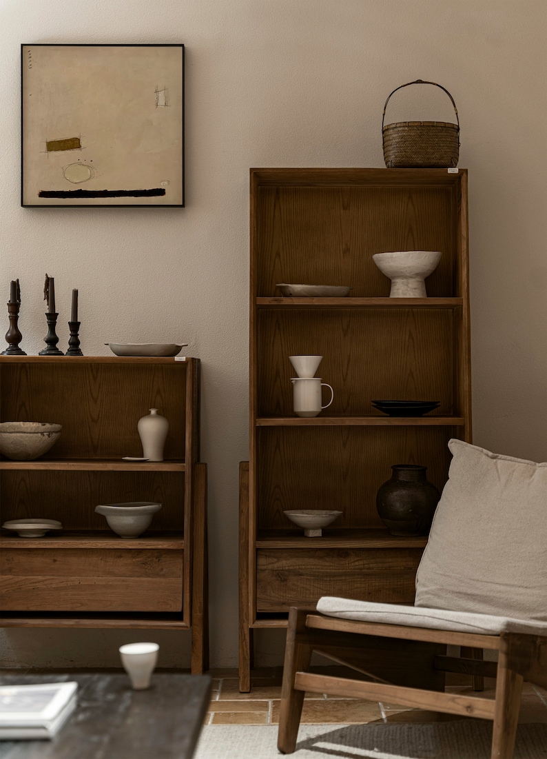 May include: Two wooden bookshelves with open shelves and drawers, filled with ceramic bowls, vases, and other decorative items. A wicker basket sits on top of one shelf. A beige armchair with a cushion sits in front of the shelves.