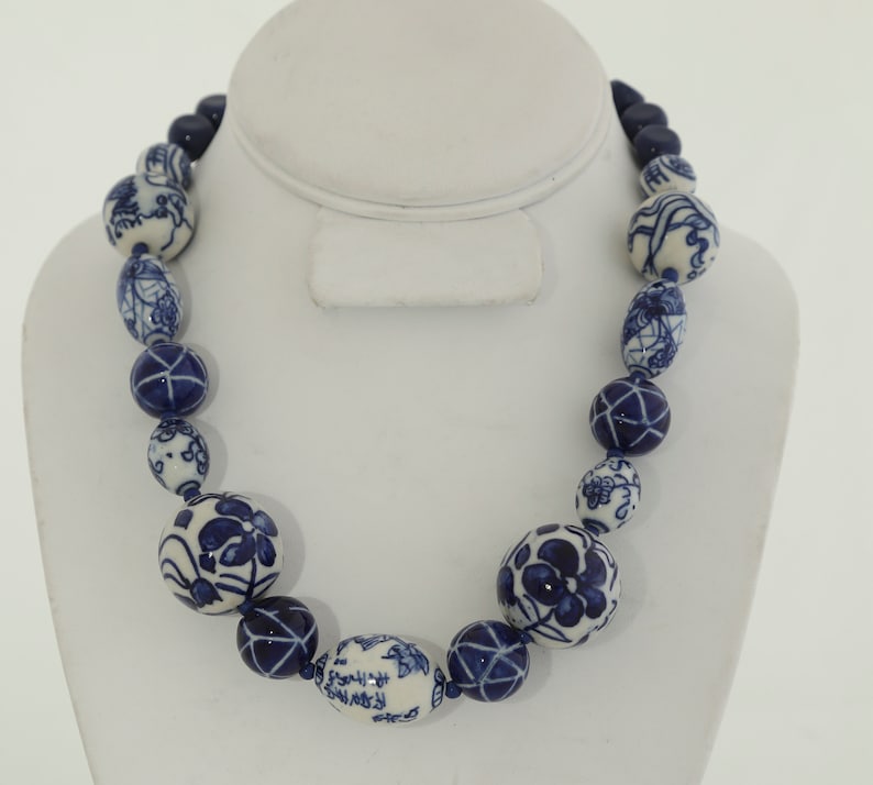 May include: A blue and white ceramic bead necklace with a variety of floral and geometric patterns. The necklace is made of large, round beads and is strung on a blue cord.