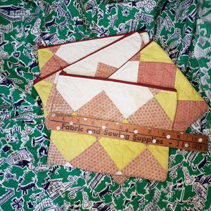May include: Three quilted fabric pouches with a yellow and brown geometric pattern. The pouches have a red zipper and are stacked on top of each other. A wooden ruler with the text "Fabric and Sewing Supplies" is lying on top of the pouches.