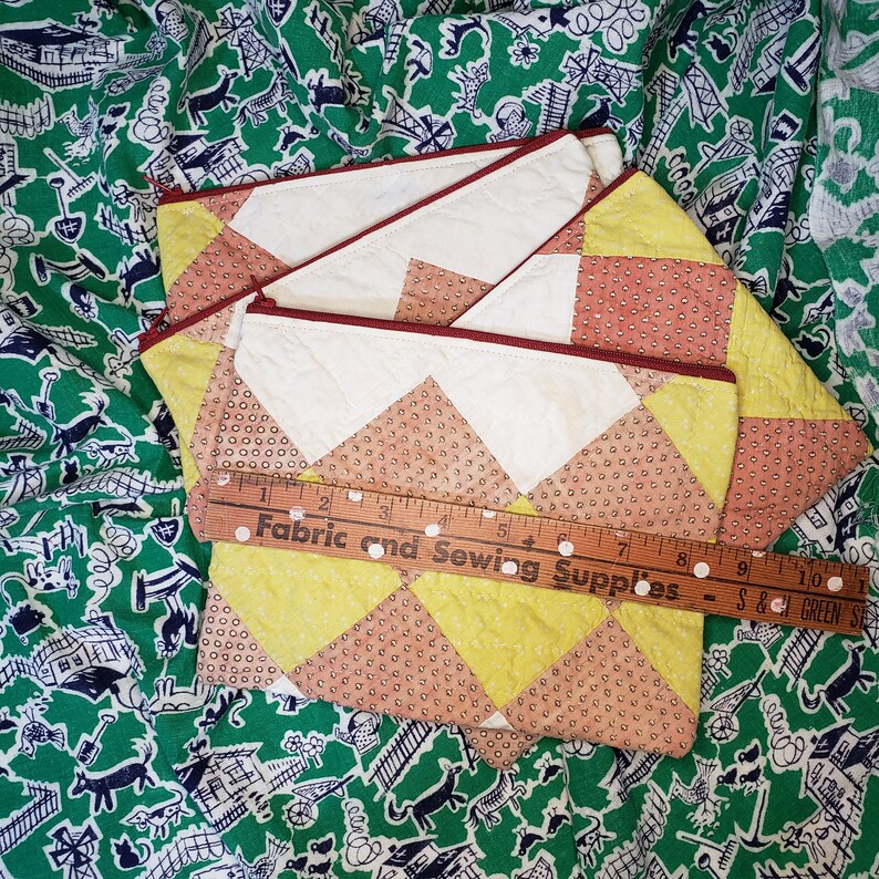 May include: Three quilted fabric pouches with a yellow and brown geometric pattern. The pouches have a red zipper and are stacked on top of each other. A wooden ruler with the text "Fabric and Sewing Supplies" is lying on top of the pouches.