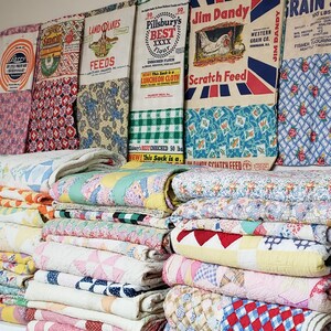 May include: A stack of vintage quilts in various colors and patterns, including pink, blue, yellow, and white. The quilts are arranged in a pile, with the top quilt folded in half. The quilts are made of different fabrics, including cotton and wool. The quilts are displayed in front of a wall with several vintage feed sacks, including Pillsbury's Best Flour, Jim Dandy Scratch Feed, and Land O'Lakes Feeds.