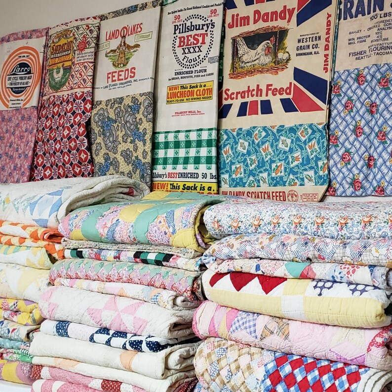 May include: A stack of vintage quilts in various colors and patterns, including pink, blue, yellow, and white. The quilts are arranged in a pile, with the top quilt folded in half. The quilts are made of different fabrics, including cotton and wool. The quilts are displayed in front of a wall with several vintage feed sacks, including Pillsbury's Best Flour, Jim Dandy Scratch Feed, and Land O'Lakes Feeds.
