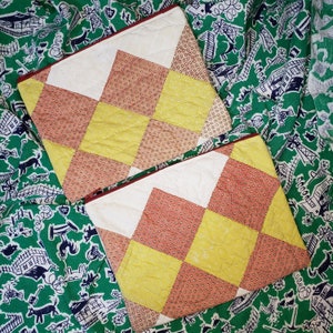 May include: Two quilted fabric squares with a checkered pattern in shades of yellow, red, and white. The squares are made of a soft, textured fabric and have a vintage aesthetic.