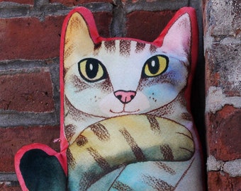 May include: A blue and gray cat-shaped plush toy with a white face and black eyes. The toy is sitting on a brick wall.