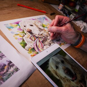 Puede incluir: Una pintura de acuarela de un perro golden retriever con una bufanda colorida alrededor del cuello. El artista est&aacute; usando un pincel para agregar detalles a la pintura.