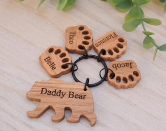 May include: A wooden keychain with a bear-shaped charm that says "Daddy Bear" and four paw-shaped charms with names: Belle, Theo, Florence, and Jacob. The keychain is on a wooden surface with green leaves.