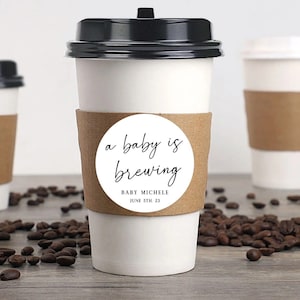 Può includere: Una tazza di caffè bianca con un manicotto marrone e un adesivo rotondo che dice "a baby is brewing" con il testo "BABY MICHELE JUNE 5TH, 23" sotto. La tazza è circondata da chicchi di caffè.