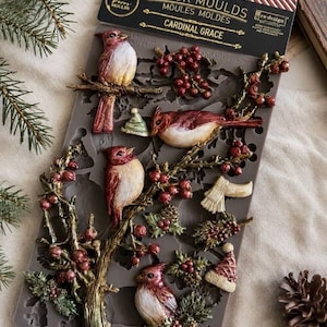 May include: A gray silicone decor mould titled Cardinal Grace by Re-Design with Prima. The mould features raised designs of three red cardinals perched on branches with berries, pinecones, a small hat, a scarf, and additional winter foliage.