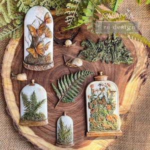 May include: A collection of seven decorative glass domes with various nature-themed scenes. The domes feature butterflies, ferns, and flowers, all rendered in intricate detail. The domes are arranged on a wooden surface with the text "CASAMIA re-design" visible in the background.