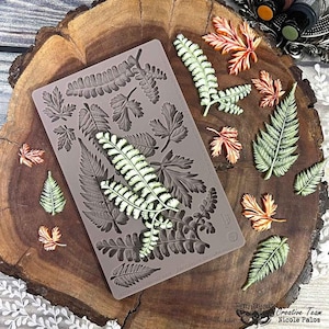 May include: A silicone mold with various leaf designs, alongside several painted leaf embellishments. The mold is a muted gray color, and the leaves are painted in shades of green, orange, and white. The items are arranged on a wooden surface.