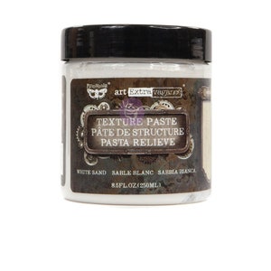 May include: A white jar of texture paste with a black lid. The label reads "Finnabair art Extra vagance Texture Paste Pâte de Structure Pasta Relieve White Sand Sable Blanc Sabbia Bianca 8.5 fl oz (250 ml)"