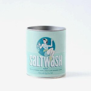 May include: A cylindrical, light teal container with a silver rim. The container features a vintage illustration of a woman and the text "SALT WASH" in a retro font. Additional text reads "FOR A LAYERED SUN & SALT AIR SOAKED LOOK Made with Real Sea Salt."