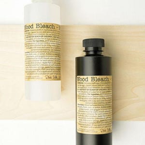 May include: Two bottles of wood bleach, one clear and one black, with white labels. The labels have black text that reads "Wood Bleach" and safety warnings. The bottles have white and black caps.