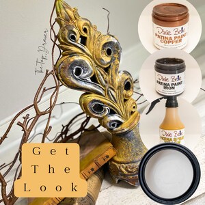 May include: A decorative metal ornament with a gold and black finish, styled with a distressed look. The ornament is displayed on a book with a wooden surface. Three bottles of Dixie Belle Patina Paint are shown in the image: Copper, Iron, and a spray bottle of Patina.