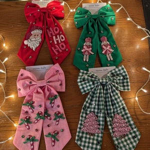 May include: Four Christmas-themed hair bows in red, green, pink, and green-and-white checkered patterns. The red bow features a Santa design and the text "HO HO HO." The green bow has two doll figures. The pink bow has candy cane designs, and the checkered bow has Christmas tree designs.