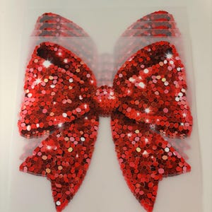 May include: A large red sparkly bow made of sequins. The bow is on a clear plastic background.