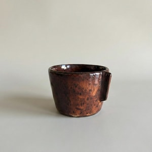 May include: A handmade, brown ceramic mug with a unique, rectangular handle. The mug has a textured surface and a rich, earthy color, perfect for coffee or tea. The mug is approximately 3 inches tall.