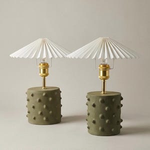 May include: A pair of table lamps featuring cylindrical olive green bases with a textured, studded surface. Each lamp has a polished brass stem and socket, topped with a white, pleated, conical paper shade that flares outward at the edges.