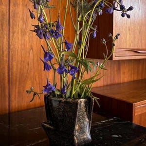 May include: A dark, geometric vase holds a vibrant arrangement of purple flowers and green stems. The vase has a textured, metallic finish. The flowers are in full bloom, creating a striking contrast against the dark vase and the wooden background.