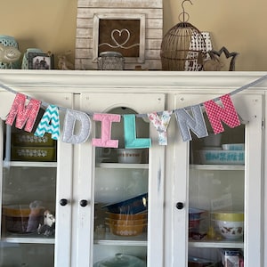 May include: A white cabinet with glass doors and a fabric banner that reads "MADILYN" in pink, gray, blue, and white letters. The banner is decorated with polka dots, zigzags, and floral patterns.