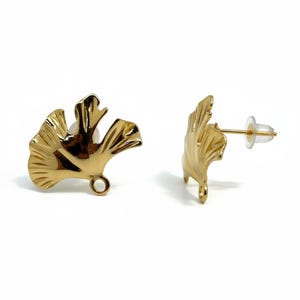May include: A pair of gold-coloured stud earrings shaped like ginkgo leaves. The earrings have a detailed, textured surface and a post with a clear backing. The earrings are displayed against a white background.