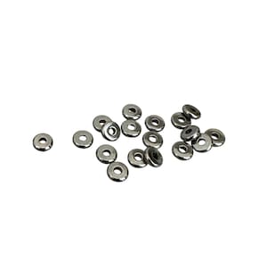 Peut inclure: Un groupe de perles d'espacement en métal argenté, dispersées sur un fond blanc. Les perles sont rondes avec un dessus et un dessous plats, et ont un petit trou au centre.