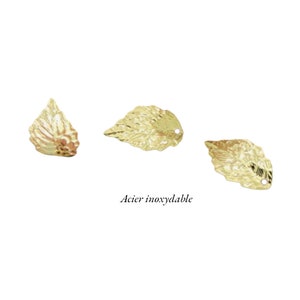 Peut inclure: Trois breloques en forme de feuille en métal doré avec une surface texturée. Les feuilles ont un trou en haut pour être attachées à des bijoux ou d'autres objets artisanaux. Le texte "Acier inoxydable" est écrit sous les feuilles.