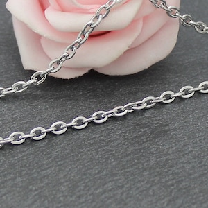 Puede incluir: Un collar de cadena de plata con un delicado dise&ntilde;o de eslabones ovalados. La cadena est&aacute; sobre una superficie gris con una rosa rosa en el fondo.
