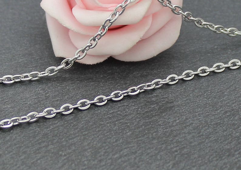 Puede incluir: Un collar de cadena de plata con un delicado dise&ntilde;o de eslabones ovalados. La cadena est&aacute; sobre una superficie gris con una rosa rosa en el fondo.