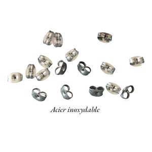 Könnte beinhalten: Nahaufnahme einer Gruppe silberner Ohrringverschlüsse, die auf einem weißen Hintergrund verstreut sind. Der Text "Acier inoxydable" ist am unteren Rand des Bildes sichtbar.