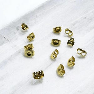 Könnte beinhalten: Nahaufnahme einer Gruppe von goldfarbenen Ohrringverschlüssen. Die Verschlüsse sind aus Metall und haben eine glatte Oberfläche. Sie sind für die Verwendung mit Steckerohrringen konzipiert.