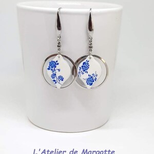 May include: A pair of silver-toned drop earrings featuring a circular metal frame surrounding a white, diamond-shaped pendant. Each pendant is decorated with a blue floral pattern. The earrings hang from silver-toned hooks. Text reads L'Atelier de Margotte.