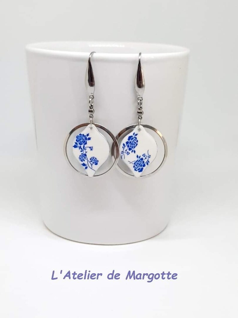 May include: A pair of silver-toned drop earrings featuring a circular metal frame surrounding a white, diamond-shaped pendant. Each pendant is decorated with a blue floral pattern. The earrings hang from silver-toned hooks. Text reads L'Atelier de Margotte.