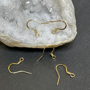 Può includere: Quattro ganci per orecchini color oro sono esposti su uno sfondo bianco cristallino e una superficie grigio scuro. I ganci sono realizzati in filo sottile, con un design curvo e una piccola spirale nella parte inferiore. I ganci sono per la creazione di gioielli.